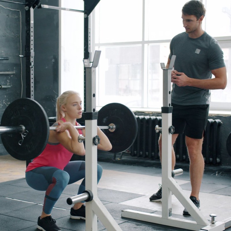 A coach standing beside a lifter squatting in a rack.